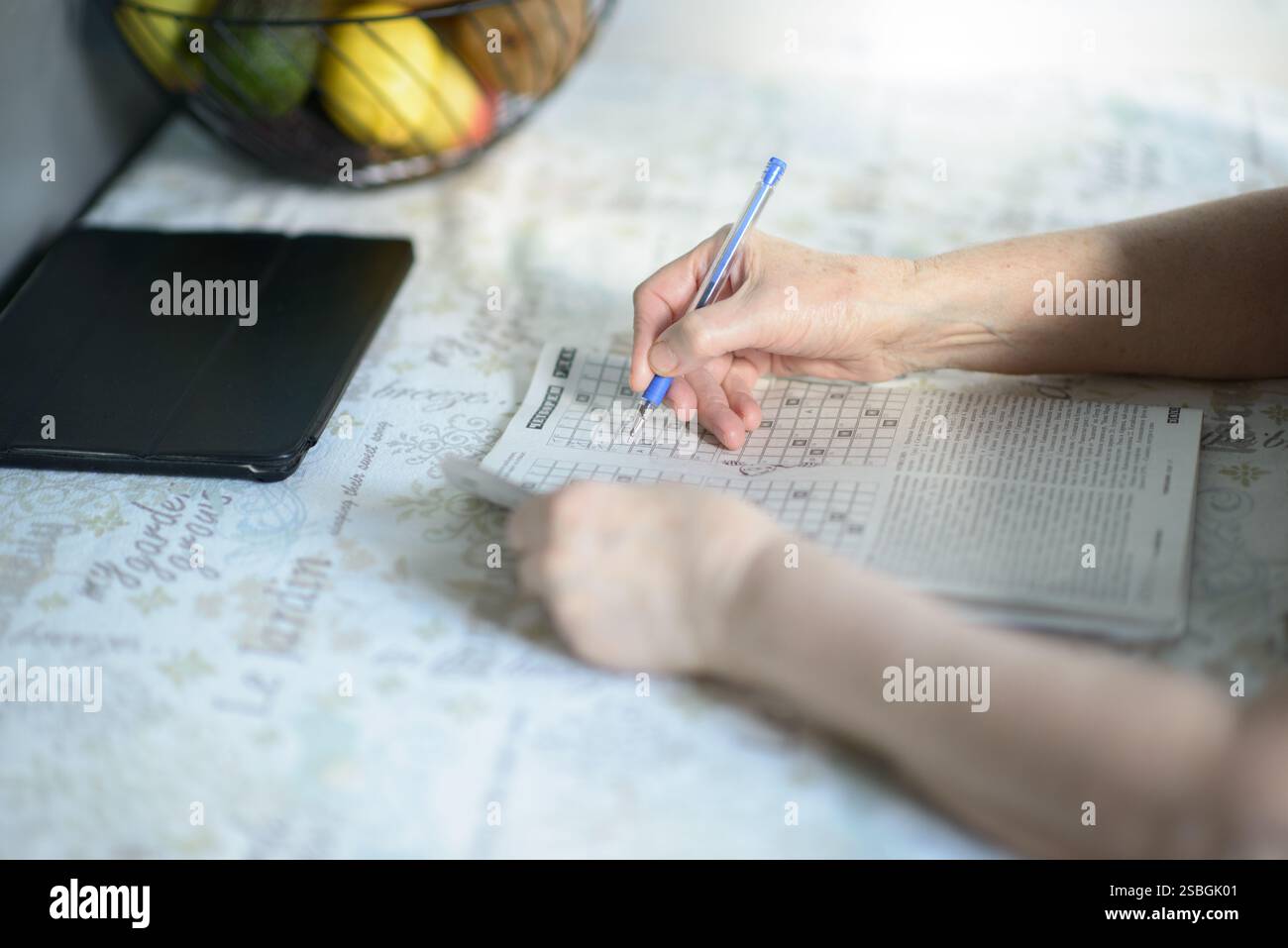 Una donna anziana che risolve un rompicapo con una penna, seduta a un tavolo con una tavoletta e una ciotola di frutta sullo sfondo. L'immagine acquisisce i concentrati Foto Stock