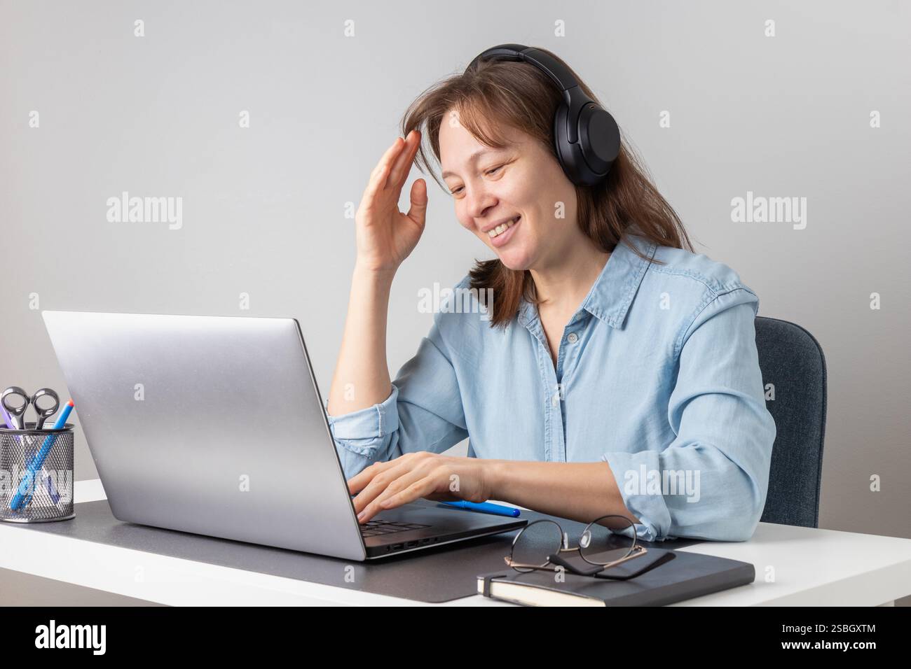 Una donna allegra e sorridente con le cuffie che utilizzano il laptop, che guarda lo schermo del PC, lavora da remoto come freelance da casa. Postura corretta, felicità Foto Stock