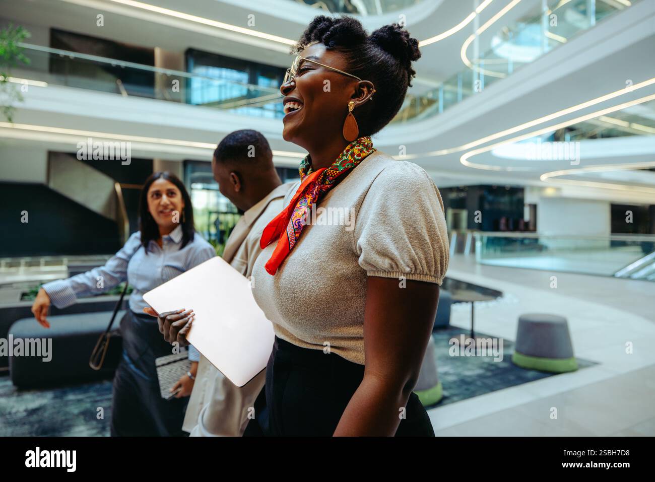 Un'allegra donna d'affari africana si impegna con i colleghi in un moderno spazio open Office. Trasuda cordialità mentre tiene in mano un laptop, simboleggiando la p Foto Stock