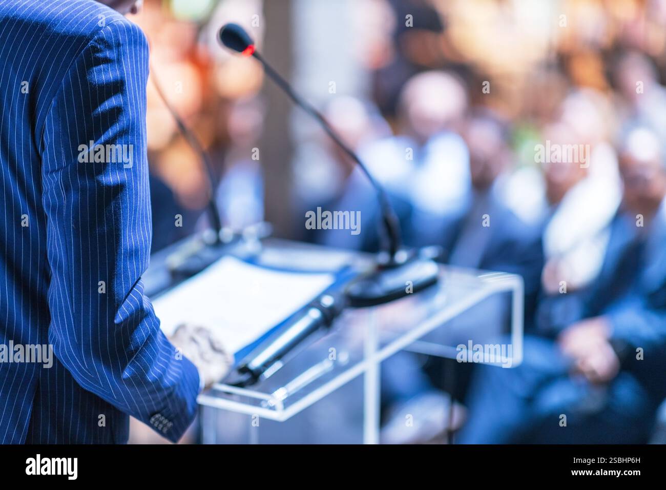 Un oratore professionista con una tuta blu si erge su un podio, offrendo una presentazione a un pubblico attento e sfocato in un ambiente formale, Foto Stock