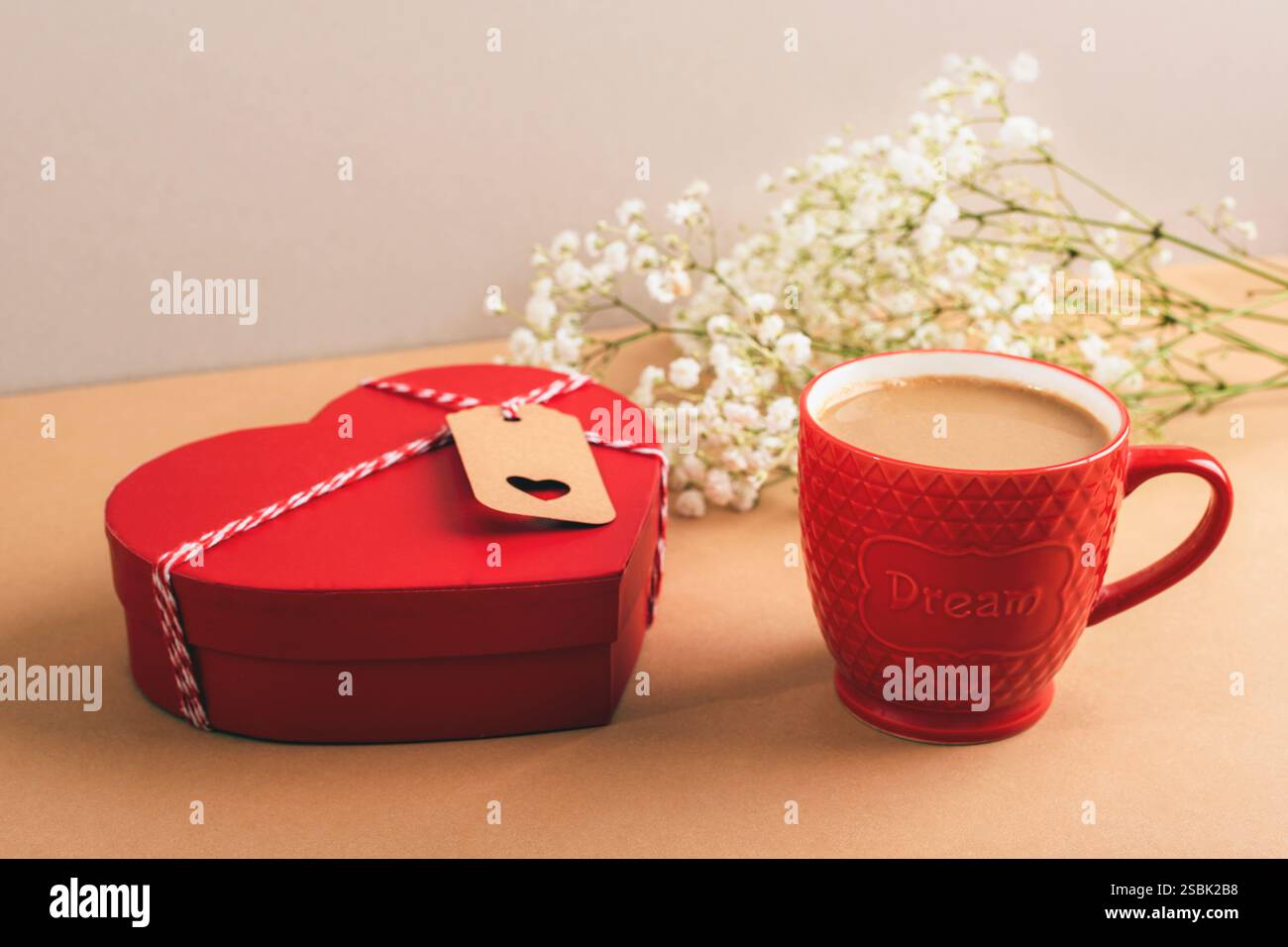 Scatola rossa a forma di cuore e tazza rossa con caffè, fiori di respiro del bambino su sfondo beige, a simboleggiare l'amore e i momenti intimi. Il giorno di San Valentino. Foto Stock