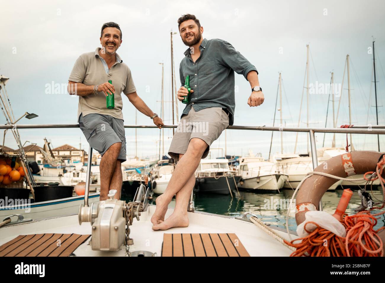 Due uomini che si rilassano su uno yacht con bevande, godendosi un momento informale al porticciolo con barche sullo sfondo, rappresentando il tempo libero e l'amicizia. Foto Stock Due uomini che si rilassano su uno yacht con bevande, godendosi un momento informale al porticciolo con barche sullo sfondo, rappresentando il tempo libero e l'amicizia. Foto Stock