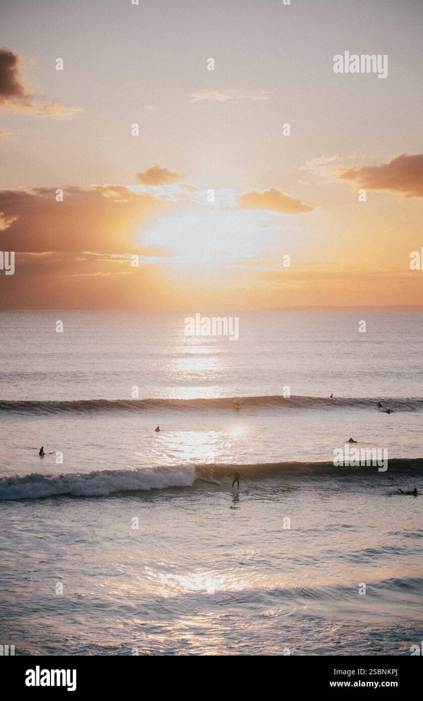Il sole tramonta sull'oceano, proiettando un caldo bagliore sull'acqua. Un gruppo di surfisti sta cavalcando le onde, godendosi il bellissimo paesaggio Foto Stock