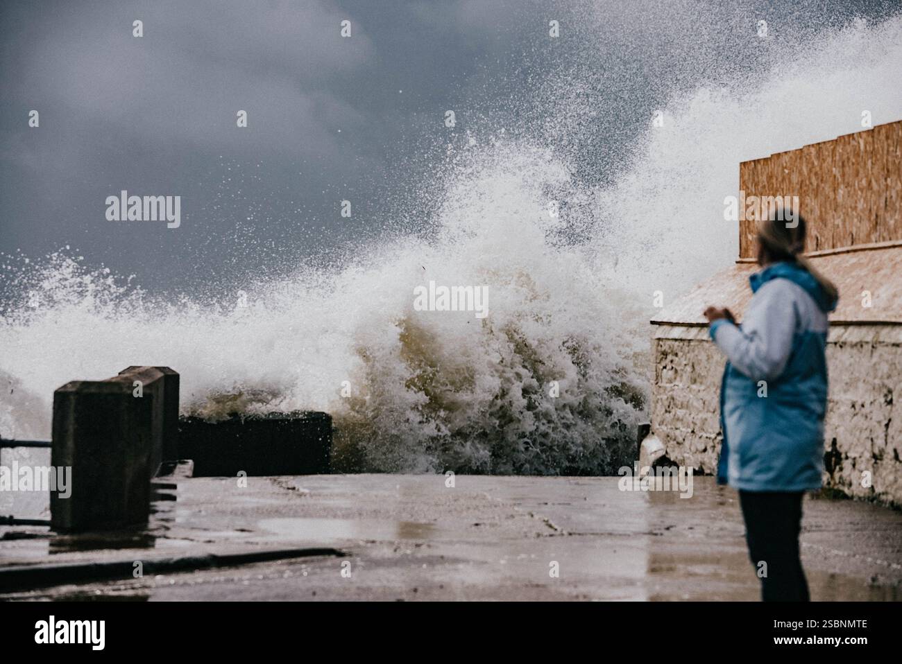 Una donna sta su un marciapiede bagnato, guardando una grande onda che si schianta sulla riva. La scena è drammatica e potente, con l'espressione della donna che si muove Foto Stock Una donna sta su un marciapiede bagnato, guardando una grande onda che si schianta sulla riva. La scena è drammatica e potente, con l'espressione della donna che si muove Foto Stock
