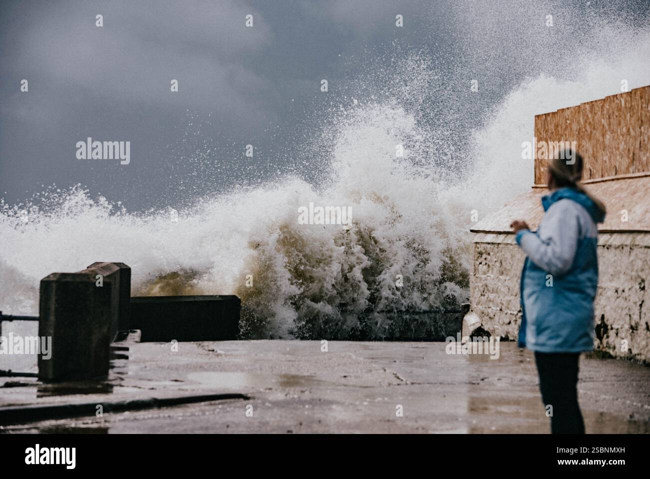 Una donna sta su un molo a guardare una grande onda che si schianta sulla riva. La scena è drammatica e potente, con l'espressione della donna che riflette l'aw Foto Stock Una donna sta su un molo a guardare una grande onda che si schianta sulla riva. La scena è drammatica e potente, con l'espressione della donna che riflette l'aw Foto Stock