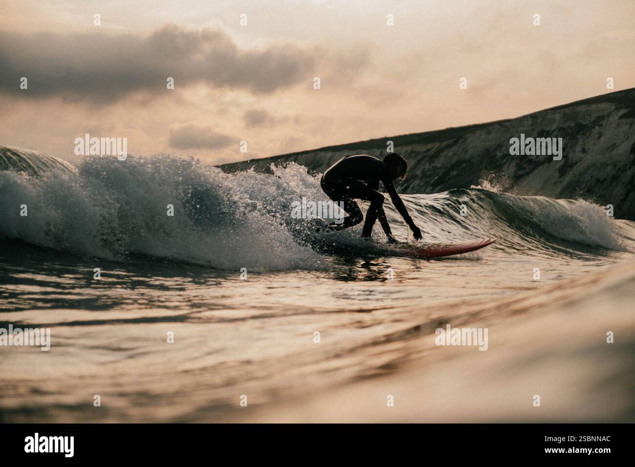 Un surfista sta cavalcando un'onda nell'oceano. Il surfista indossa una muta nera e si appoggia in avanti sulla tavola da surf. L'acqua è instabile, e la sk Foto Stock