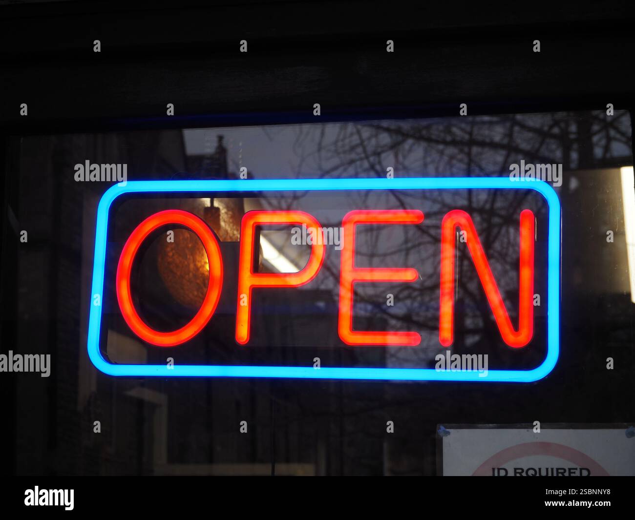 Insegna rossa al neon illuminata con cornice al neon blu nella porta del negozio di vetro con tubi fluorescenti bianchi dietro e riflesso di alberi ed edifici. Foto Stock