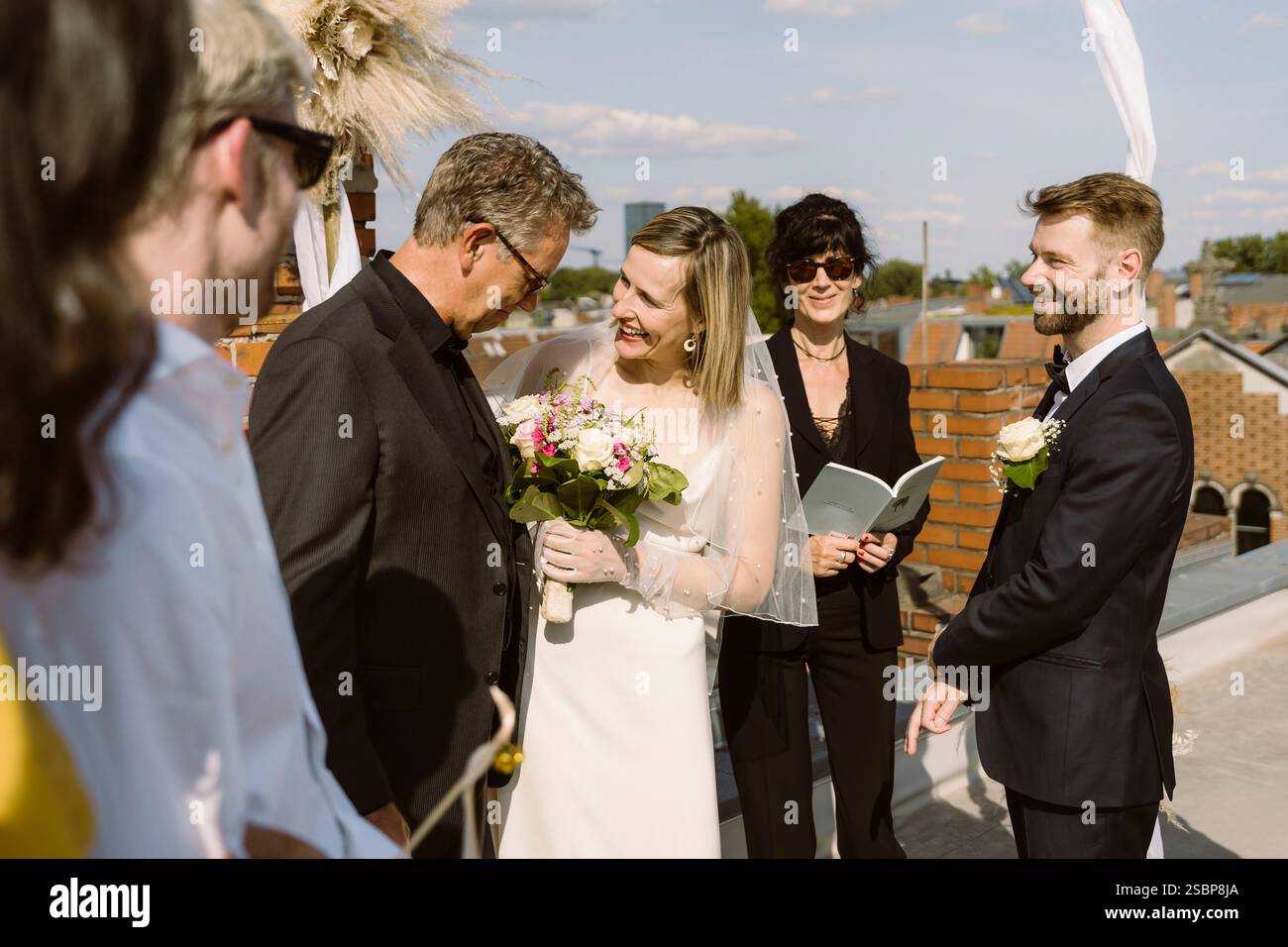 Felice sposa nuziale che tiene il bouquet e guarda il padre durante la celebrazione del matrimonio sul tetto Foto Stock