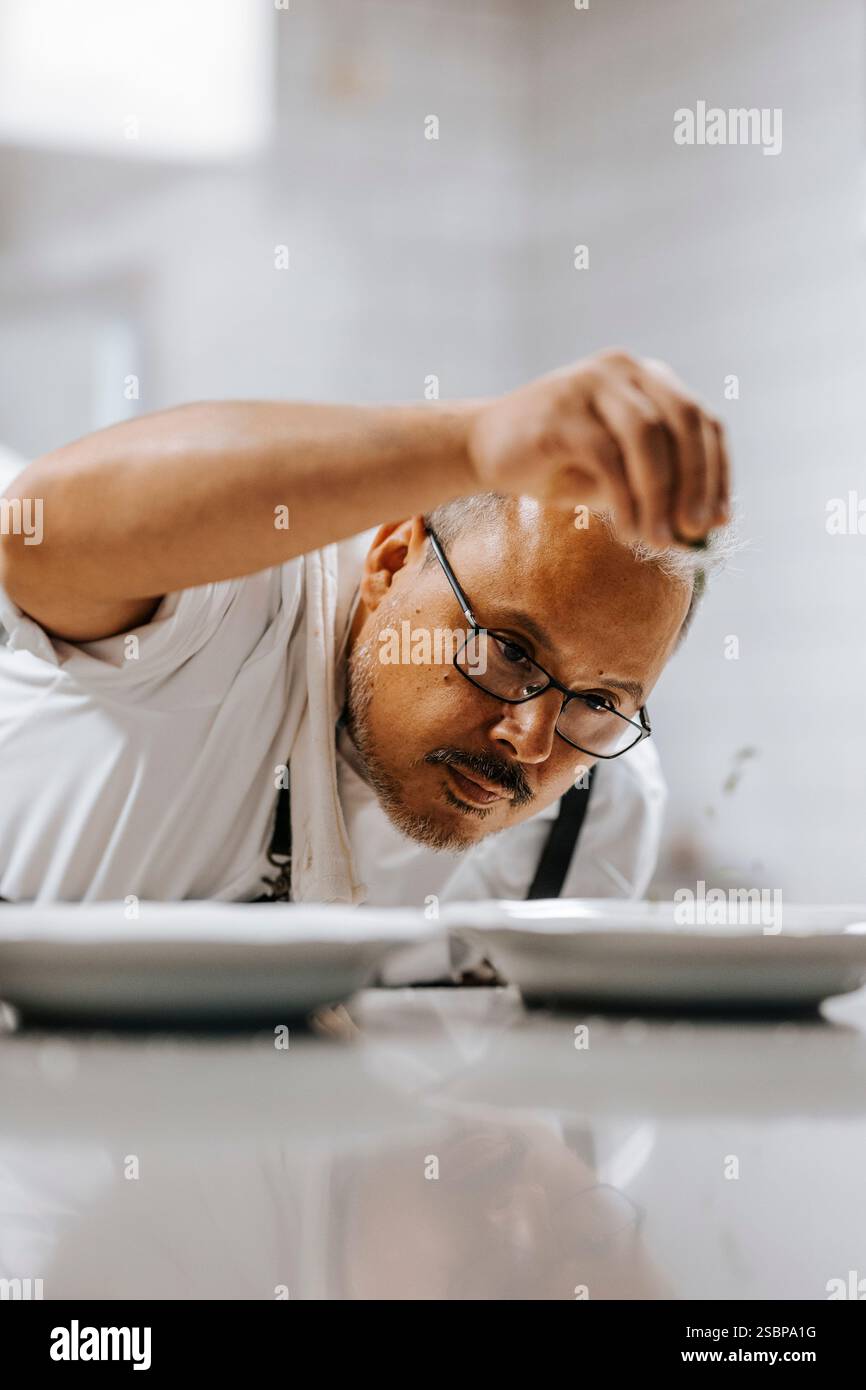 Chef maschile maturo concentrato in occhiali da vista per guarnire il cibo mentre lavora in cucina commerciale Foto Stock