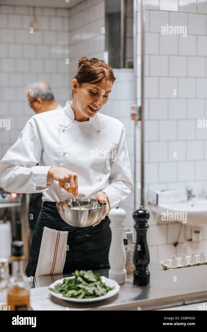 Una cuoca sorridente che mescola il cibo nella ciotola mentre si trova vicino al bancone della cucina del ristorante Foto Stock