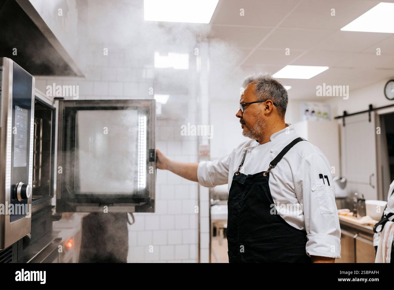 Chef maschio maturo che apre il forno mentre si trova in una cucina commerciale Foto Stock
