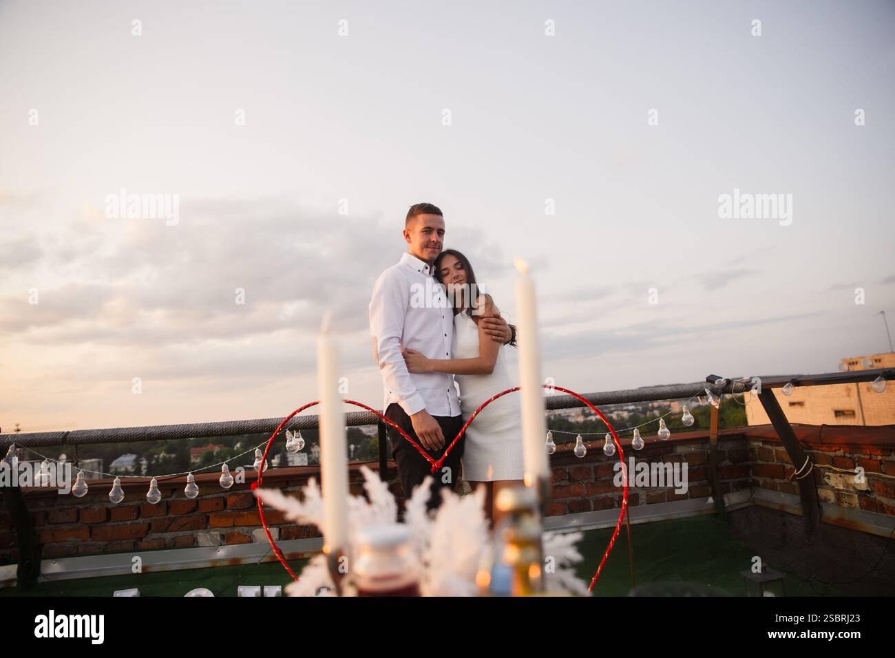 Una coppia abbraccia un tetto adornato di decorazioni mentre il sole tramonta, creando un'atmosfera pittoresca e romantica. L'illuminazione soffusa migliora l'ingresso Foto Stock
