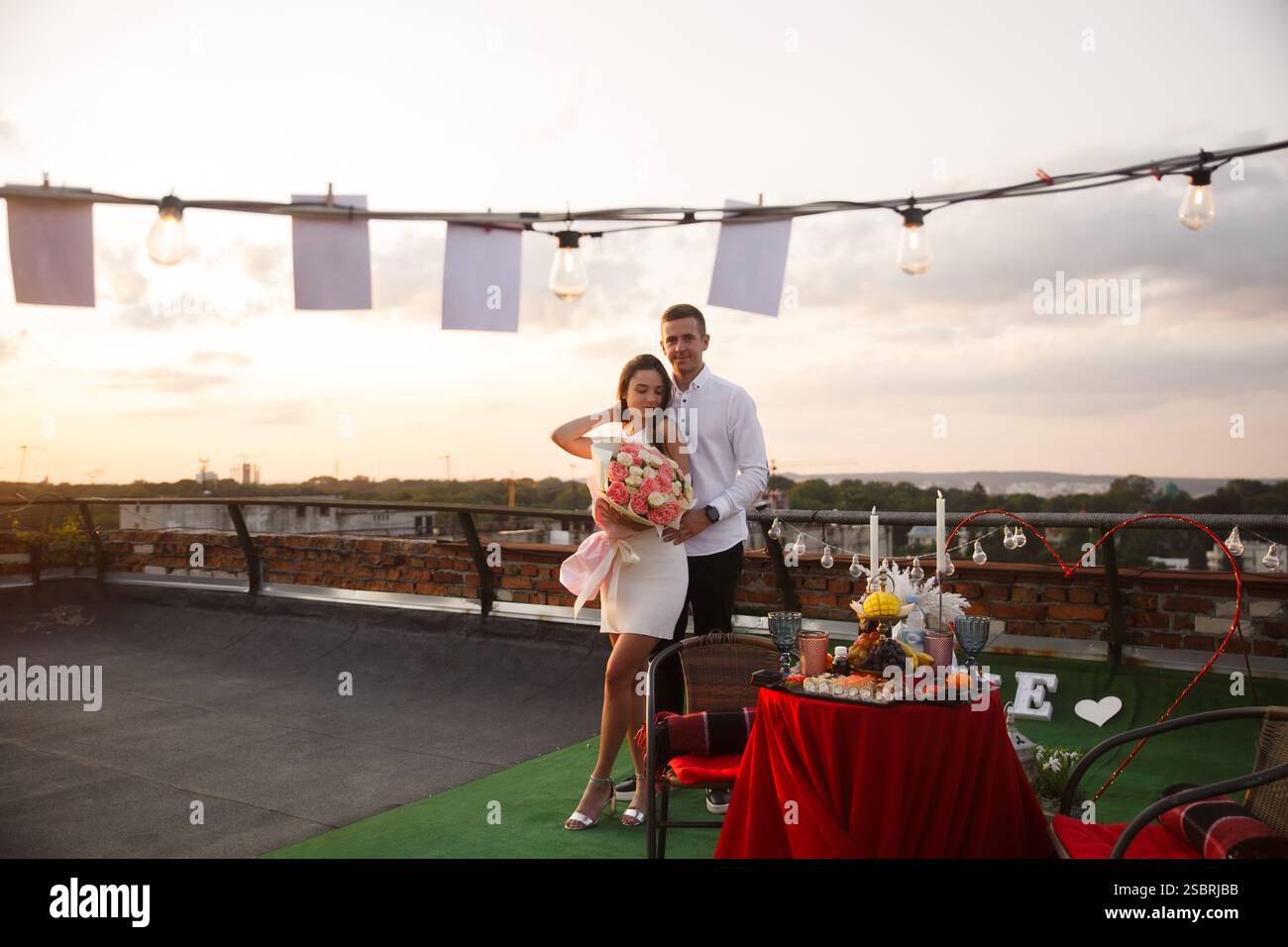 Una coppia gode di una romantica cena sul tetto durante il tramonto. L'ambientazione è caratterizzata da splendide decorazioni, un tavolo pieno di cibo e un luccicante cordoncino Foto Stock
