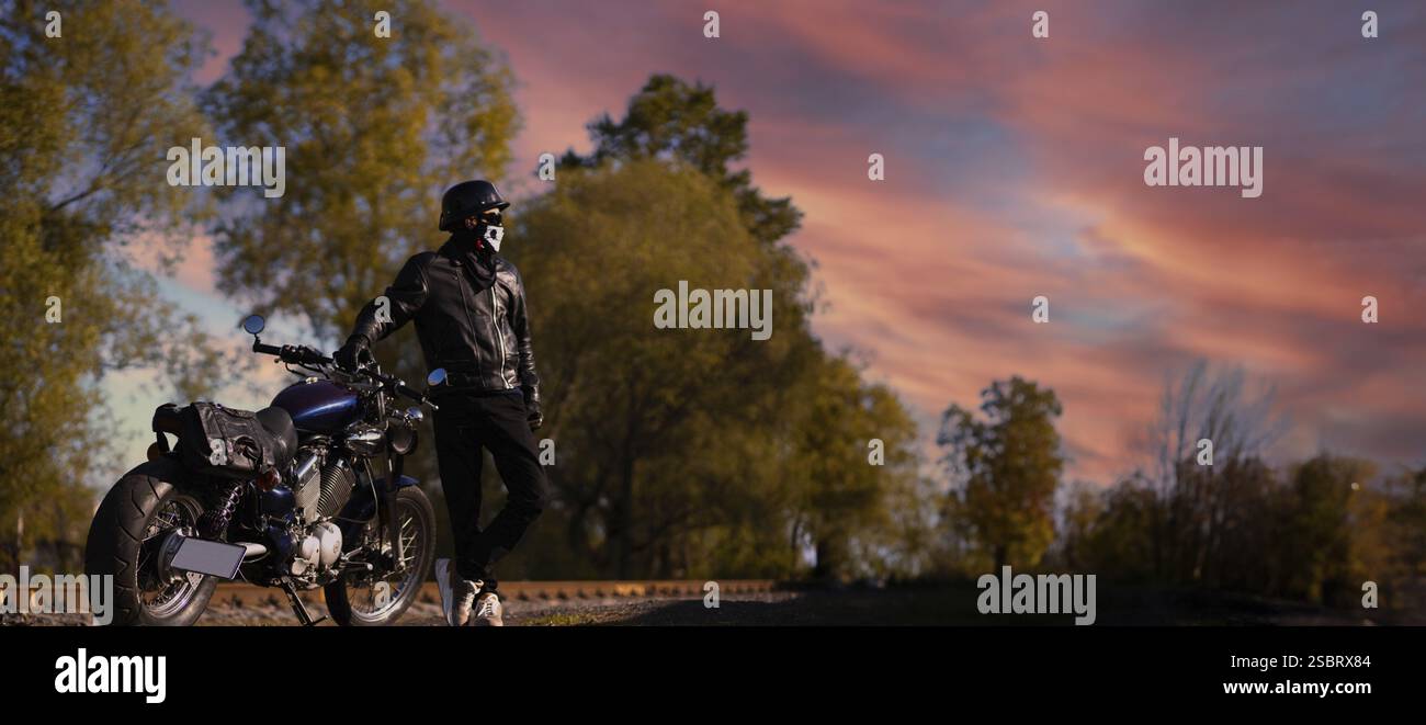 L'uomo in una giacca di pelle si trova accanto alla moto sotto un tramonto colorato, creando una vista panoramica Foto Stock L'uomo in una giacca di pelle si trova accanto alla moto sotto un tramonto colorato, creando una vista panoramica Foto Stock