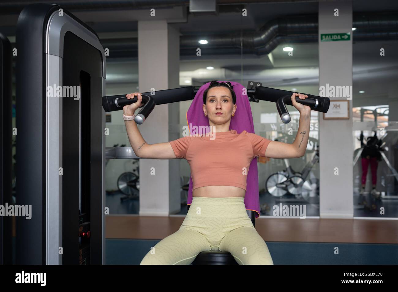 La giovane donna con un asciugamano intorno al collo esegue un esercizio di pressione sulle spalle su una macchina per il fitness, dimostrando forza e dedizione al suo workou Foto Stock