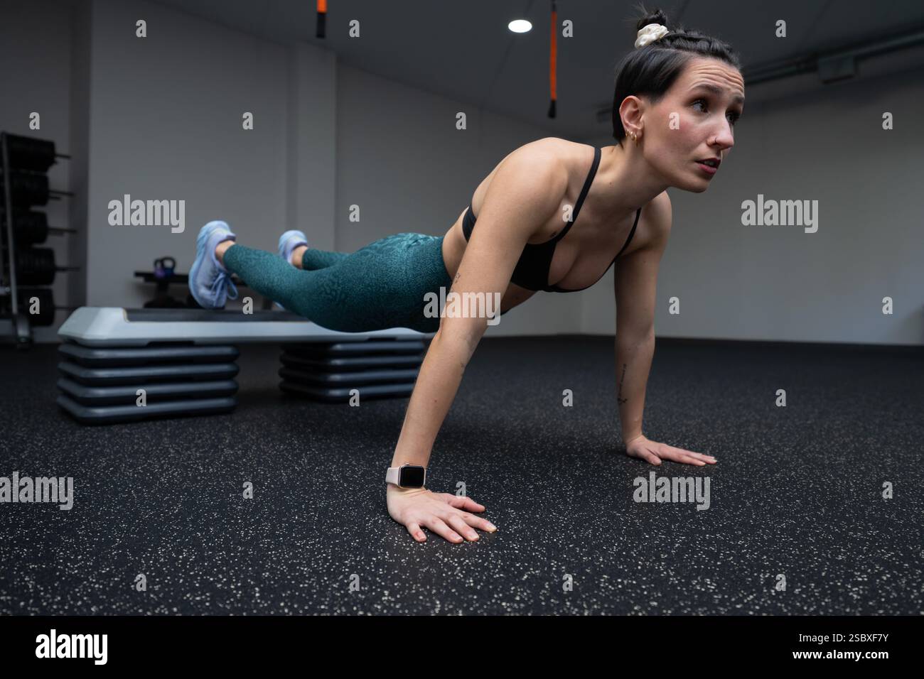 Giovane donna che esegue push up con i piedi sollevati su uno stepper aerobico in palestra, mostrando forza e fitness mentre è impegnata in un intenso allenamento Foto Stock