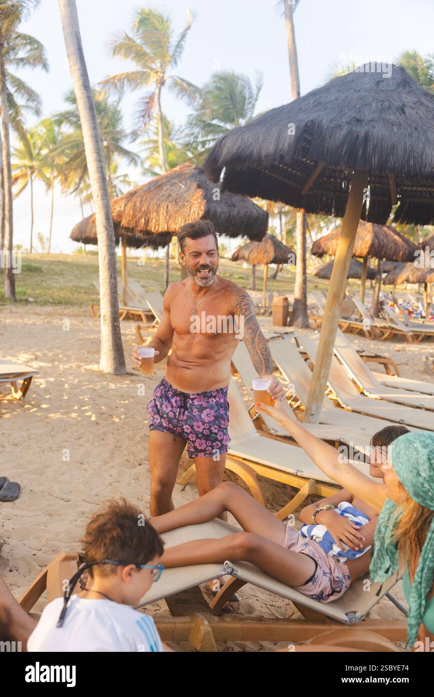 Turista di mezza età che serve un bicchiere di birra a una donna rilassata nel resort sulla spiaggia Grand Palladium Imbassai, Salvador de Bahia, Brasile Foto Stock