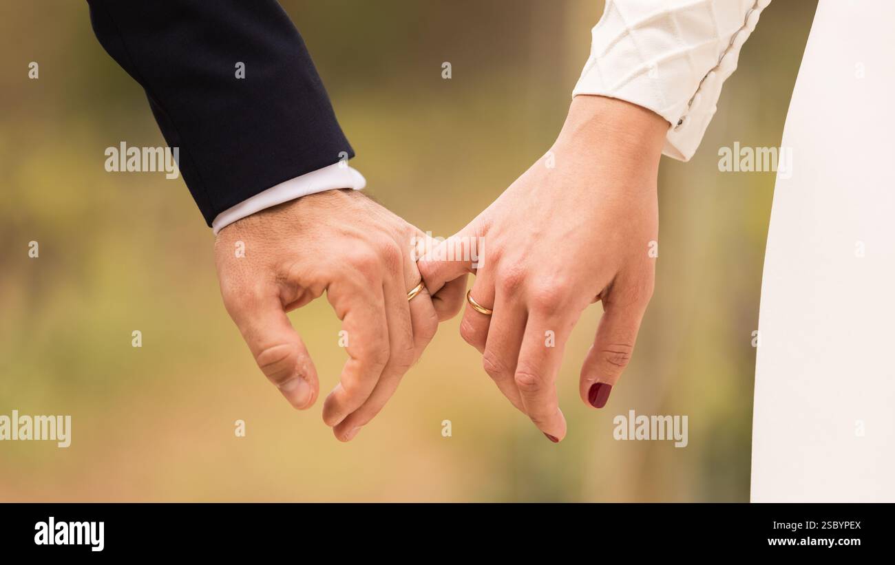 Le mani di una coppia sposata, tenendosi per mano con un piccolo dito, con anelli di marito e moglie, sfondo sfocato, abito da sposa bianco Foto Stock