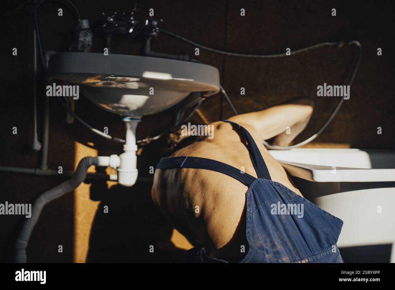 Un uomo in tuta si appoggia sotto un lavandino per lavorare sui collegamenti idraulici Foto Stock