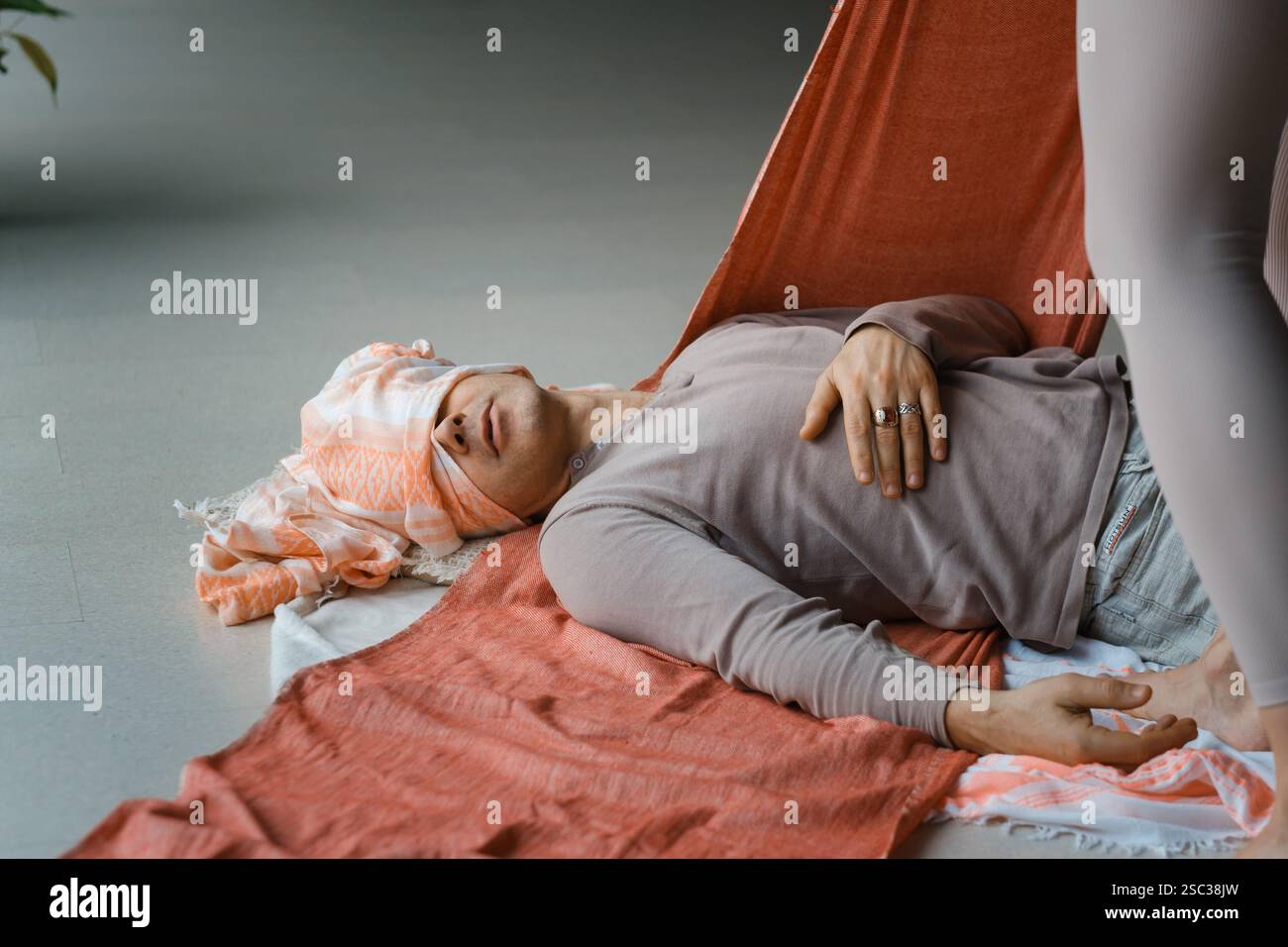 Un tranquillo ambiente interno è caratterizzato da una persona sdraiata su un tappeto, sostenuta da un tessuto morbido. Un altro individuo regola delicatamente il tessuto, aiutando in relax Foto Stock