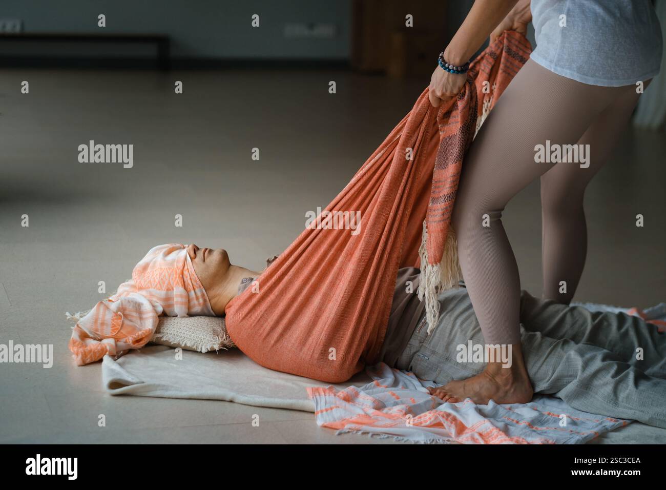 Un tranquillo ambiente interno è caratterizzato da una persona sdraiata su un tappeto, sostenuta da un tessuto morbido. Un altro individuo regola delicatamente il tessuto, aiutando in relax Foto Stock