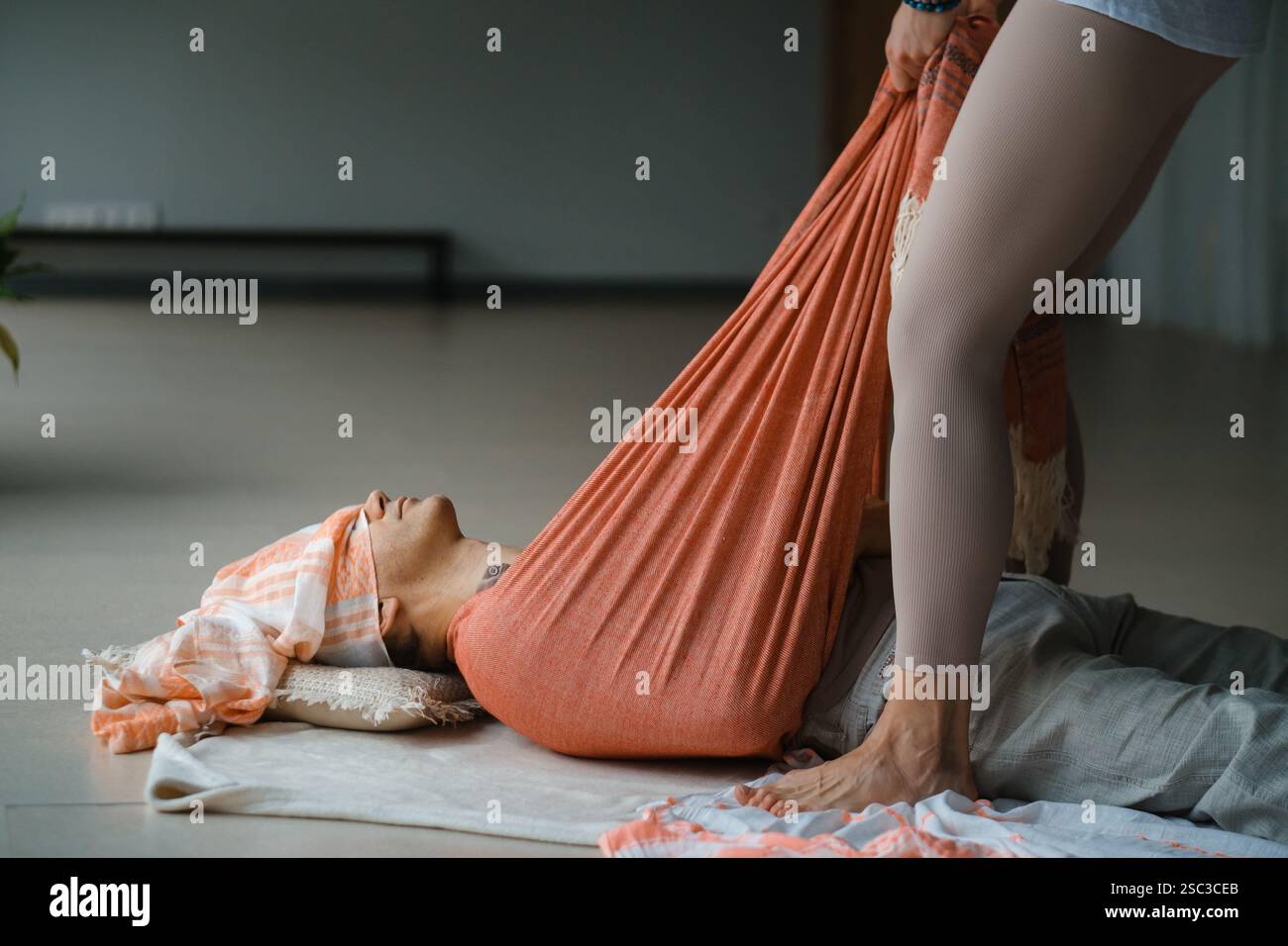 Un tranquillo ambiente interno è caratterizzato da una persona sdraiata su un tappeto, sostenuta da un tessuto morbido. Un altro individuo regola delicatamente il tessuto, aiutando in relax Foto Stock