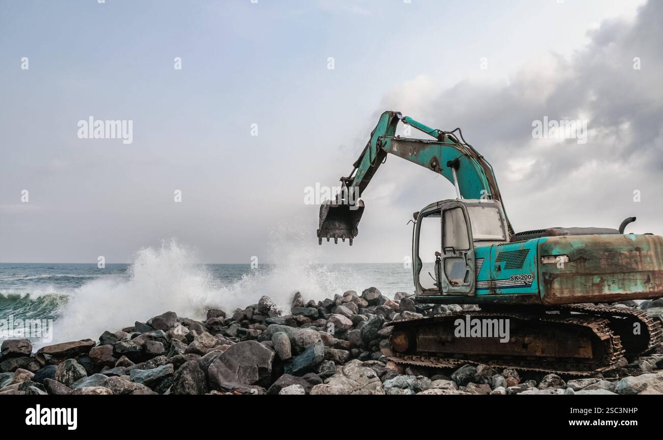 Un escavatore arrugginito di color verde acqua scava attivamente attraverso la costa rocciosa mentre le onde dell'oceano si scontrano contro la macchina, creando una dinamica scena costiera Foto Stock