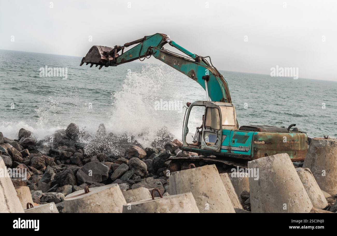 Un arrugginito escavatore di tè scava abilmente nella costa rocciosa mentre le onde dell'oceano si infrangono davanti, mostrando un cantiere in attività Foto Stock