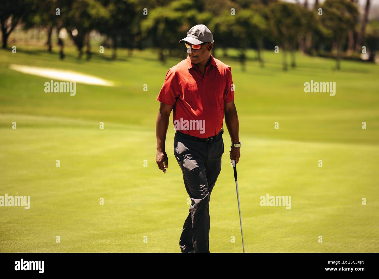Il golfista maschile cammina sul lussureggiante fairway tenendo in mano un club, indossando occhiali da sole, vicino a un bunker di sabbia, impegnato in una partita competitiva. Abbigliamento professionale per golfisti Foto Stock