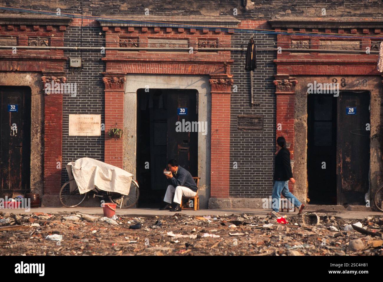 Persone di fronte a edifici in mattoni in una strada in Cina, con alcuni detriti visibili sul terreno. Foto Stock
