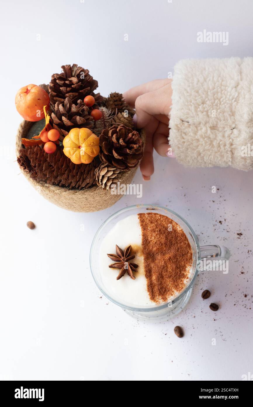 Accogliente ambiente autunnale con una mano in un morbido maglione che tocca una decorazione autunnale di coni di pino, frutti di bosco e una mini zucca accanto a un latte di spezie. Foto Stock