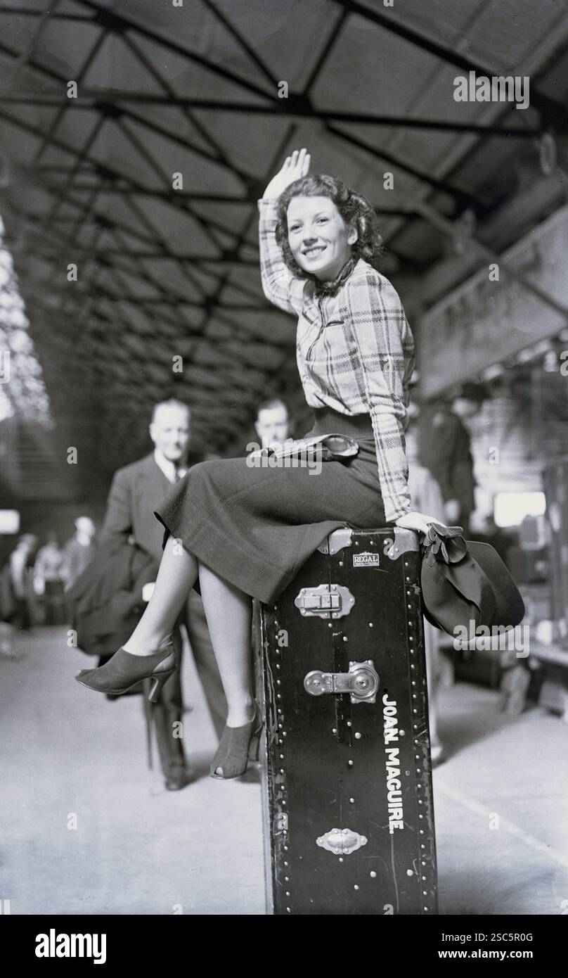 1938, storico, appena arrivato..... L'attrice americana Joan McGuire saluta mentre siede sulla sua valigia "Regal" incorniciata in legno dell'epoca su una piattaforma della stazione ferroviaria di Southampton, Inghilterra, Regno Unito. La stazione ferroviaria era vicino ai moli e serviva il porto di Southampton fino al 1966. Foto Stock
