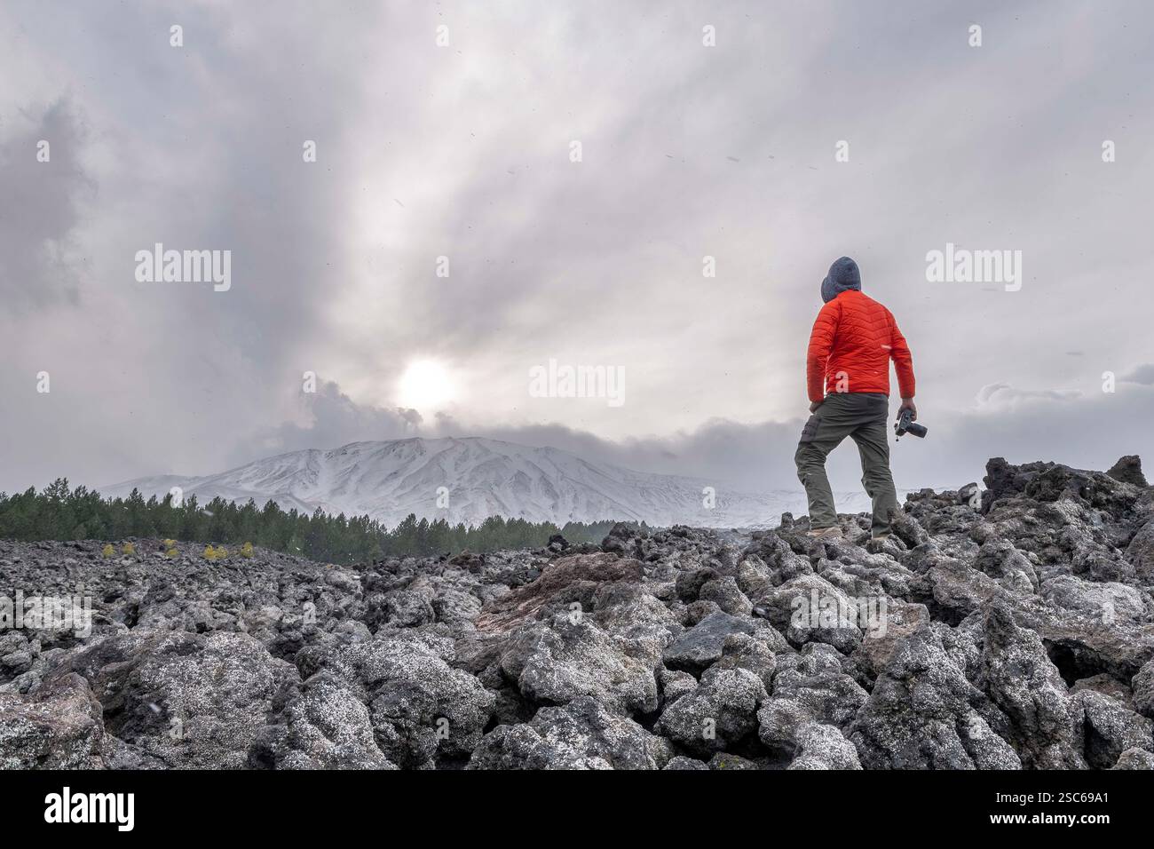 Uomo con fotocamera in piedi su un grande campo lavico dell'Etna con vista sul cratere sommitale, sul vulcano patrimonio dell'umanità dell'UNESCO, sul vulcano Stratocaster, sull'Etna e sull'Ital Foto Stock