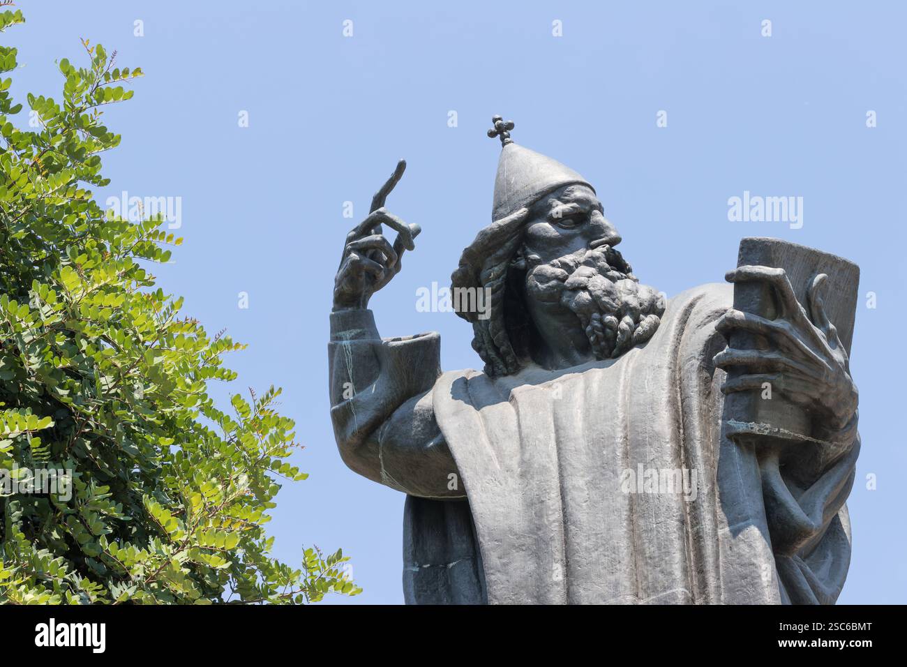 Un ambiente tranquillo caratterizzato da un campanile storico accanto a un'alta statua, circondato da una vegetazione lussureggiante, sotto un cielo azzurro e limpido, che evoca cultura e storia Foto Stock
