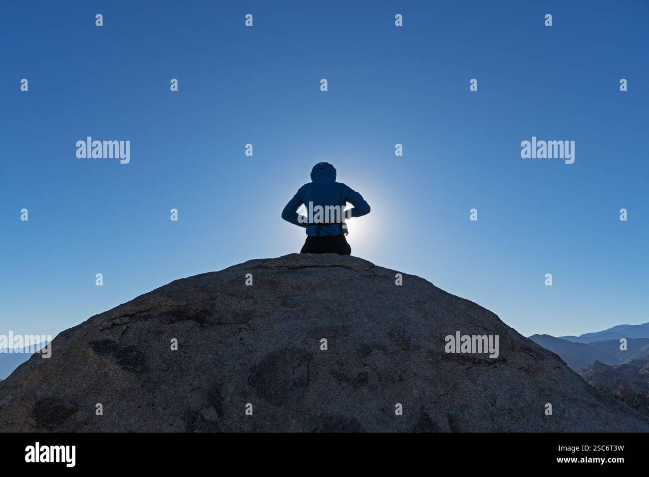 vista posteriore di una donna retroilluminata seduta sul masso in cima alla montagna con sfondo blu del cielo Foto Stock