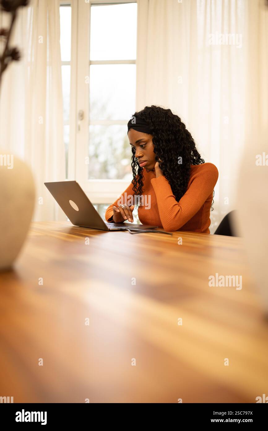 Giovane donna seduta su un tavolo di legno in un accogliente ufficio domestico, concentrata sul lavoro mentre utilizza un notebook. Indossando un maglione arancione e un cerchietto, She exu Foto Stock