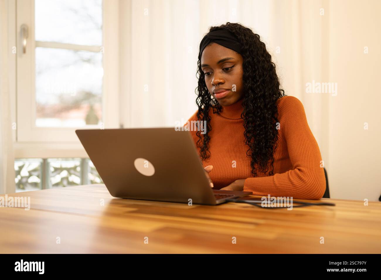 La giovane donna è seduta a un tavolo di legno vicino alla finestra con vista invernale e lavora su un laptop, indossando un maglione dolcevita arancione e un cerchietto nero Foto Stock