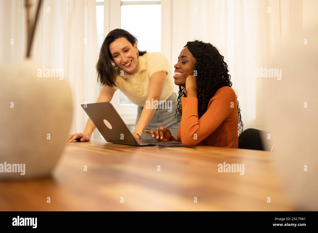Due donne imprenditrici felici lavorano insieme nel loro luminoso ufficio domestico, utilizzando un laptop e godendosi un momento di collaborazione e condivisione Foto Stock
