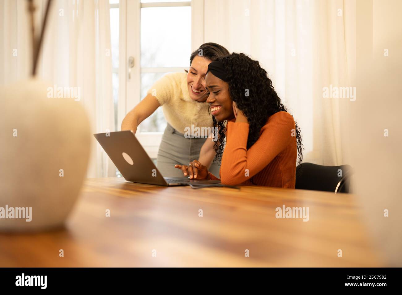 Due colleghi sorridenti stanno collaborando a un progetto, utilizzando un computer portatile su un tavolo di legno in un ufficio domestico illuminato, godendosi la loro produttività Foto Stock
