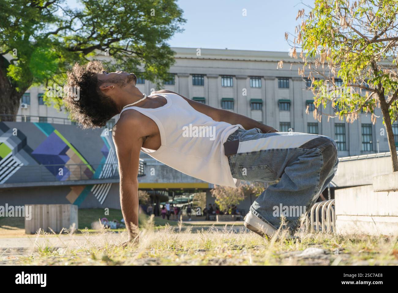 Un bel giovane latino con uno stile urbano moderno seduto sull'erba e sulla pietra in una piazza. Foto Stock