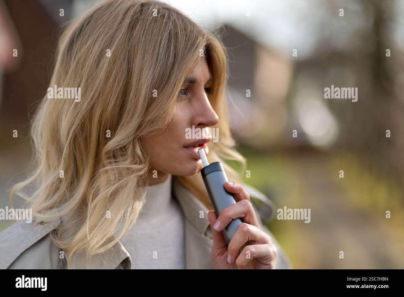 Una giovane donna con lunghi capelli biondi è all'aperto, utilizza un vaporizzatore personale mentre si impegna in una contemplazione attenta. L'impostazione è naturale Foto Stock