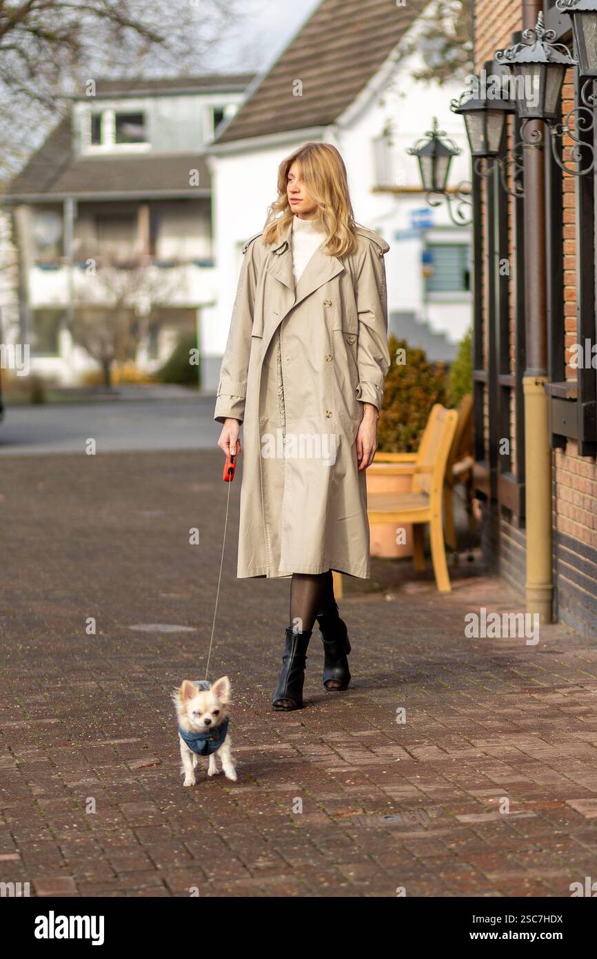 Una donna alla moda con un leggero trench passeggia lungo un sentiero di mattoni con il suo piccolo cane. Il quartiere presenta case accoglienti e un cielo limpido, indi Foto Stock
