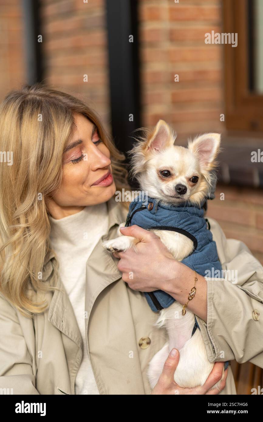 Una donna sorride calorosamente tenendo vicino il suo piccolo cane, entrambi a proprio agio nelle giornate soleggiate. L'edificio in mattoni crea un accogliente backdro Foto Stock