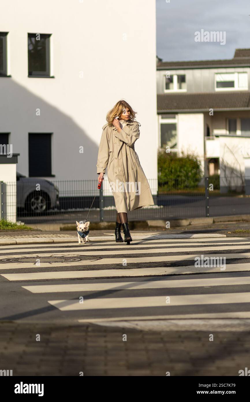 Una donna che indossa un lungo cappotto passeggia tranquillamente con il suo piccolo cane al guinzaglio mentre attraversa la strada. Il sole mette in risalto gli edifici moderni nelle vicinanze. Foto Stock
