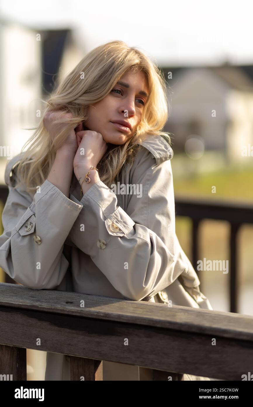 Una giovane donna dai capelli biondi si appoggia contro una ringhiera di legno, persa nel pensiero. Indossa un cappotto leggero e gode di un momento tranquillo in un vicino tranquillo Foto Stock