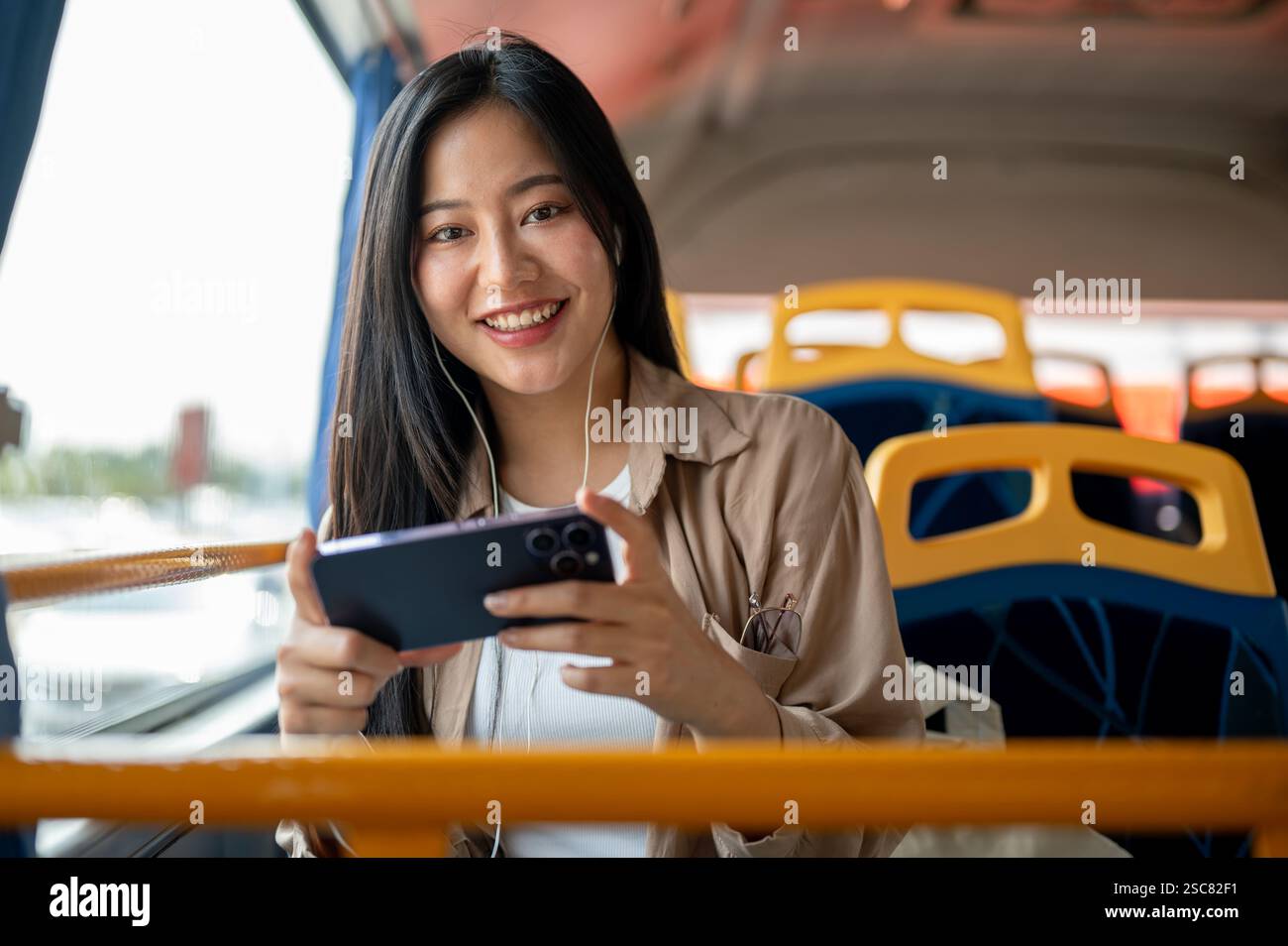 Una bella e allegra donna asiatica in abiti casual ama guardare un film o un video sul suo smartphone mentre siede davanti al finestrino su un autobus Foto Stock