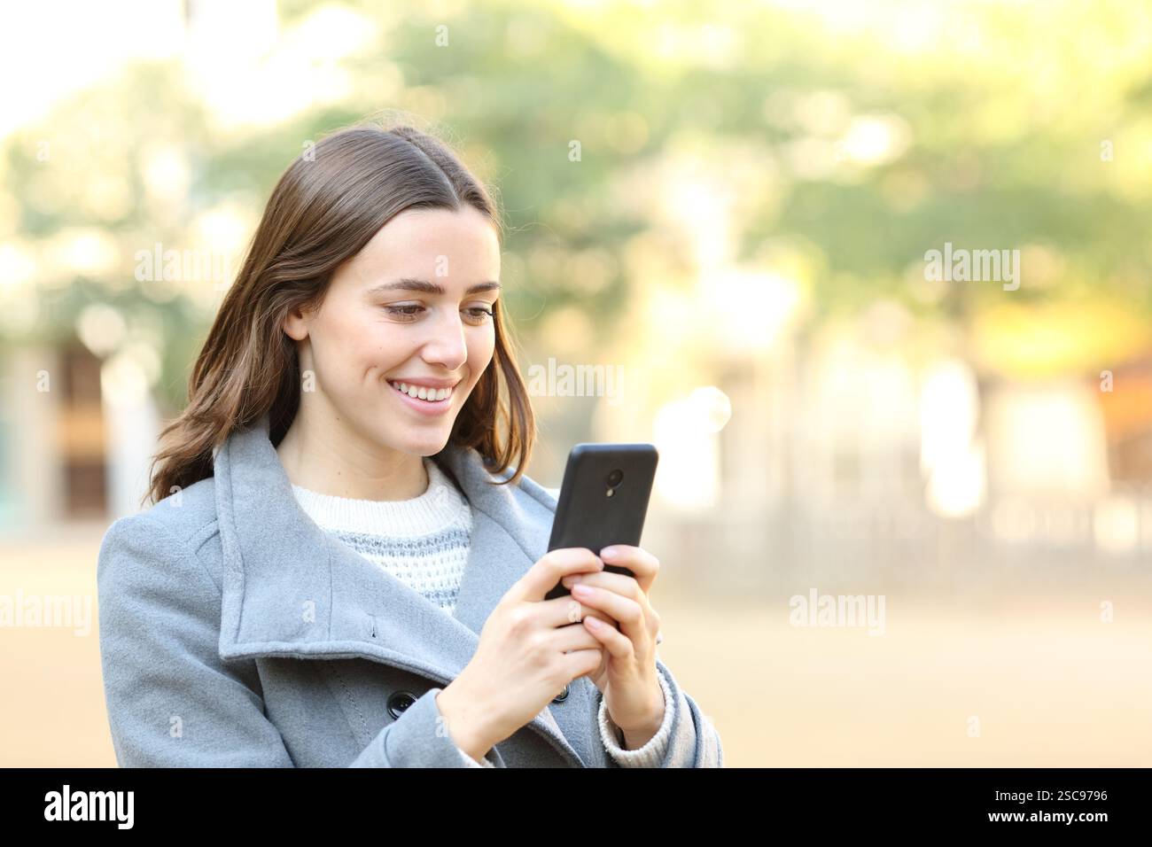 Donna felice in inverno che cammina in strada o parcheggia usando il cellulare Foto Stock