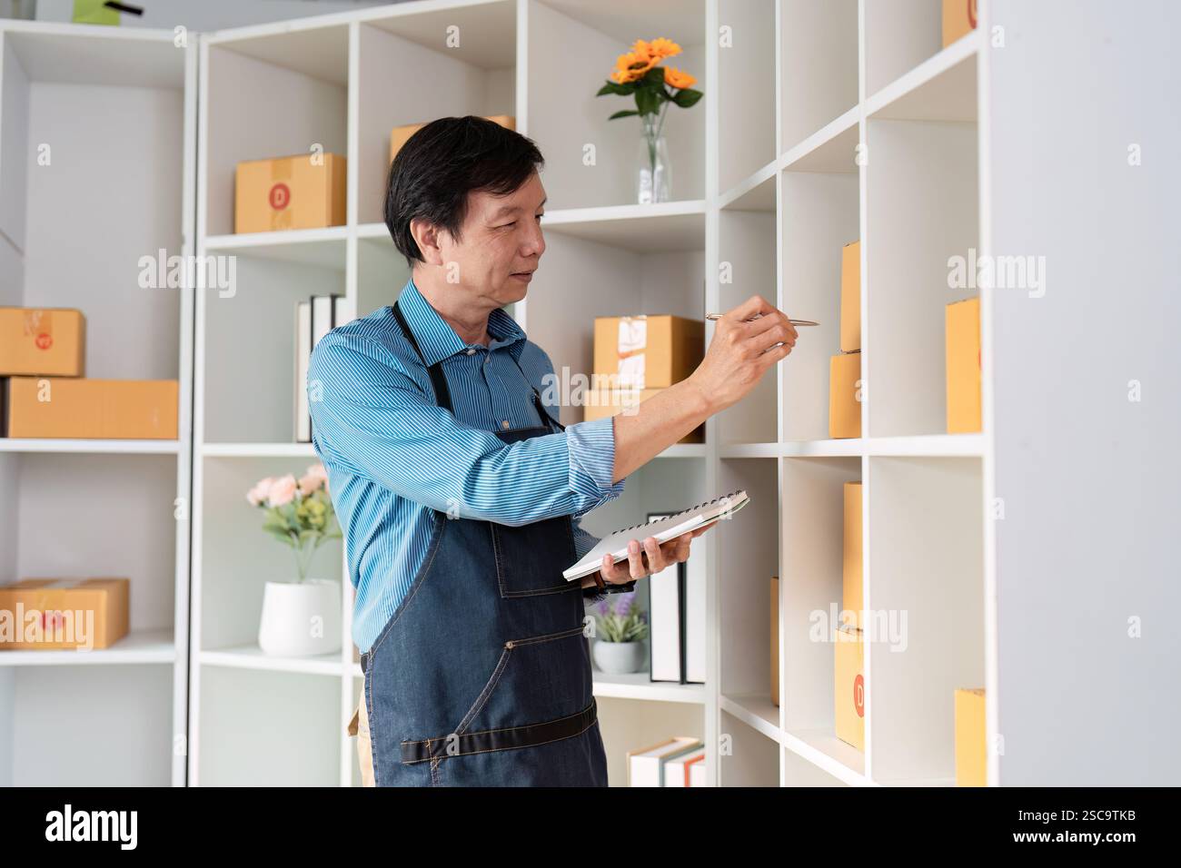 Imprenditore di e-commerce che organizza l'inventario sugli scaffali e prende appunti in un moderno spazio di lavoro pieno di scatole per la spedizione. Foto Stock