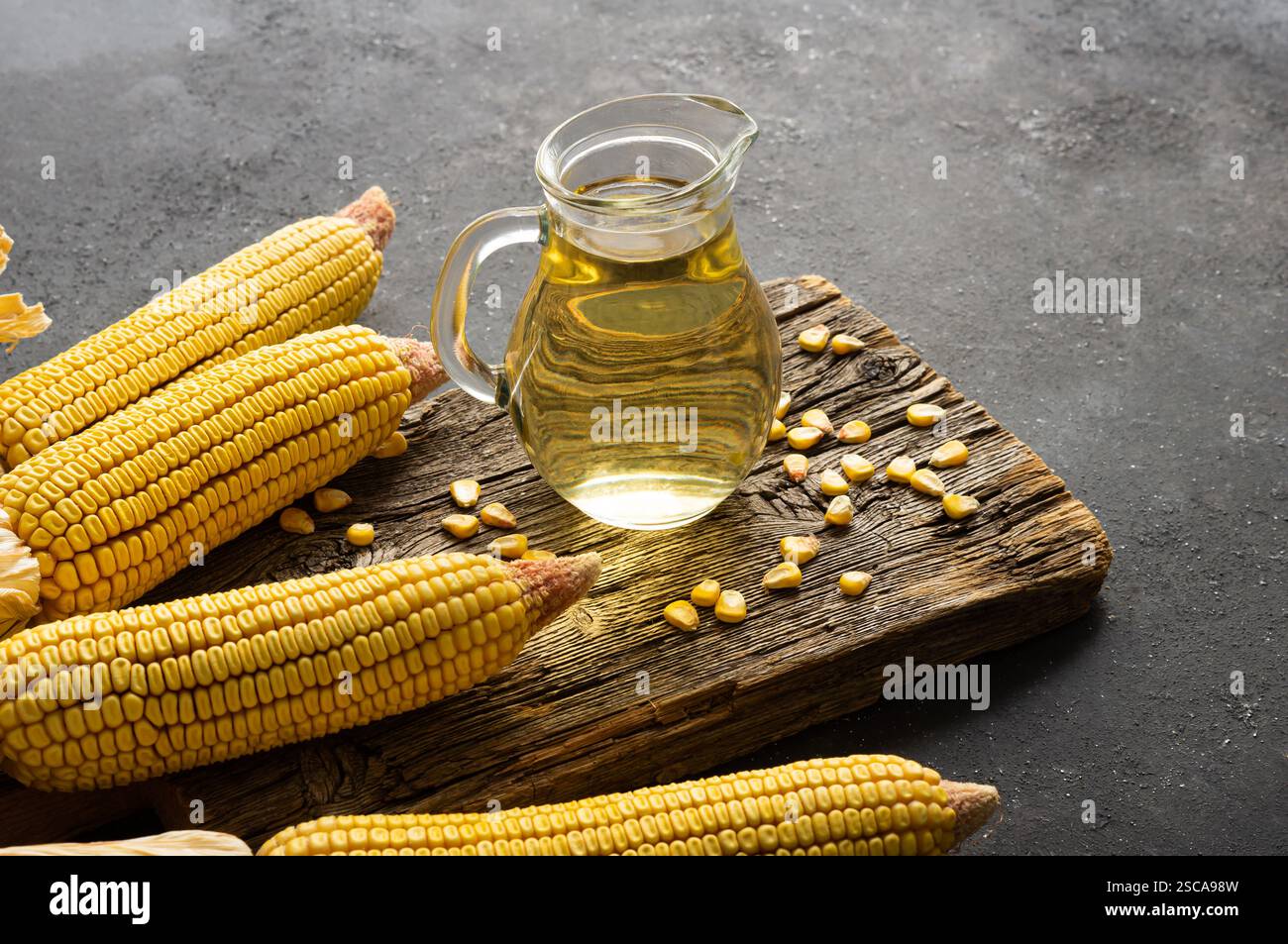 Olio di mais in un recipiente di vetro con pannocchie di mais maturi essiccate su un tavolo rustico scuro, composizione dell'olio di cottura Foto Stock