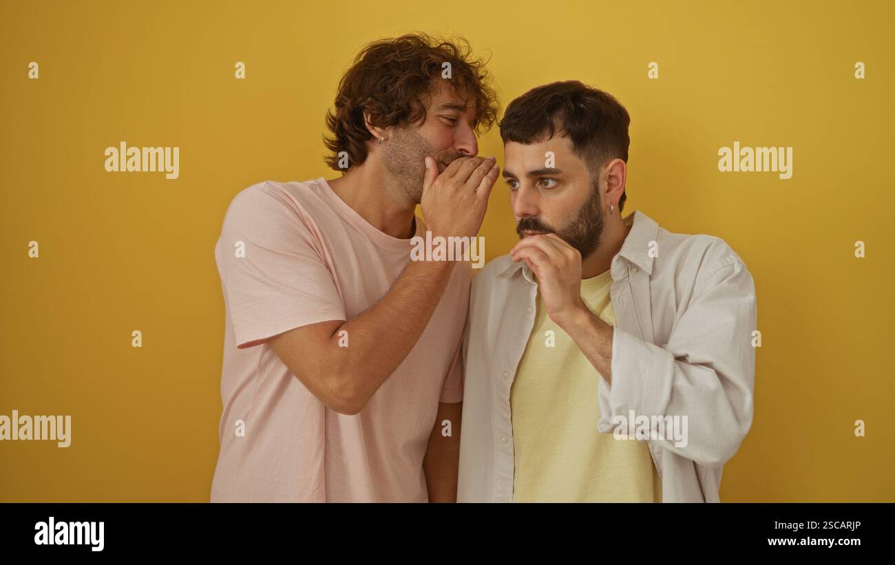 Uomini, amici, insieme, che sussurrano segreti contro un muro giallo vibrante suggeriscono una relazione forte e intima tra due fratelli o mem di famiglia Foto Stock